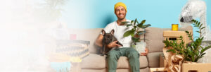 young guy sitting on a couch surrounded by all his stuff in a new apartment and holding a pug and a potted plant