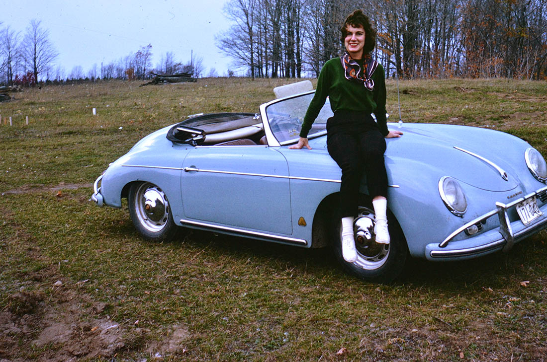 Photo of June Phelps sitting on the hood of a vintage Porsche