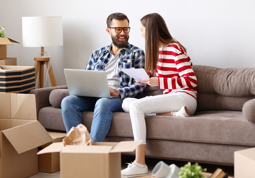 Happy young couple sitting on a couch discussing insurance amid open and partially unpacked moving boxes