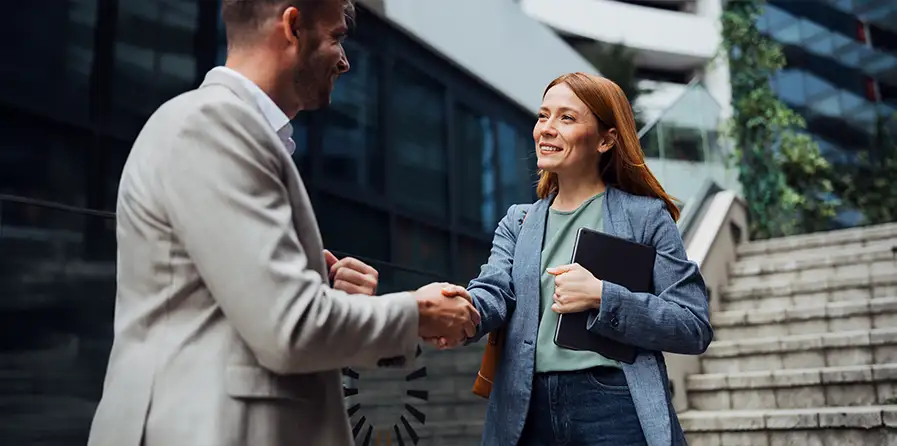 Professional businessman and businesswoman in formal attire shaking hands outdoors in front of a modern glass office building